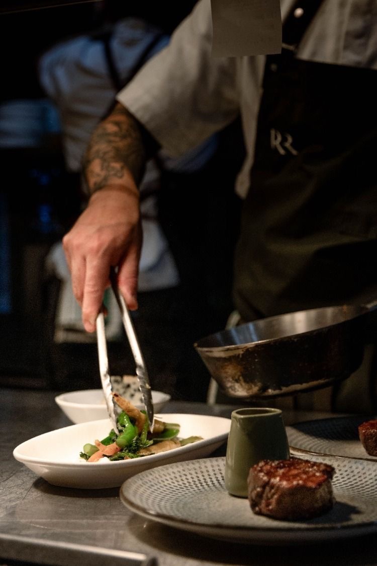 Chef plating vegetables beside a steak on the kitchen pass